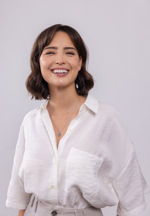 Chemise blanche texturée à boutons avec une poche poitrine, portée avec un collier en argent dont le pendentif et un pantalon gris clair.