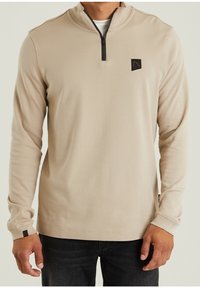 Beige Langarmpullover mit einem Halb-Reißverschluss und kontrastierendem schwarzen Reißverschluss. Mit kleinem Logo auf der Brust und gerippten Bündchen. Einfaches Design.