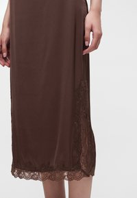 Jupe midi marron avec un liseré en dentelle à l’ourlet et sur le côté, portée par une personne debout, les mains détendues sur les côtés.
