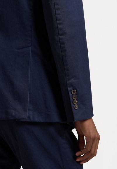 Tengerészkék blézer sima textúrával, egyetlen középen nyílt hátul, és öt barna gombbal az ujjak végén. Testre szabott fazon, struktúrált vállakkal.