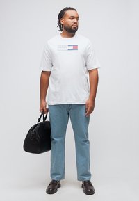 Άνδρας με ντρέντλοκς που φοράει λευκό T-shirt Tommy Hilfiger και μπλε τζιν παντελόνι, κρατώντας μαύρη δερμάτινη τσάντα duffel, στέκεται σε απλό φόντο.