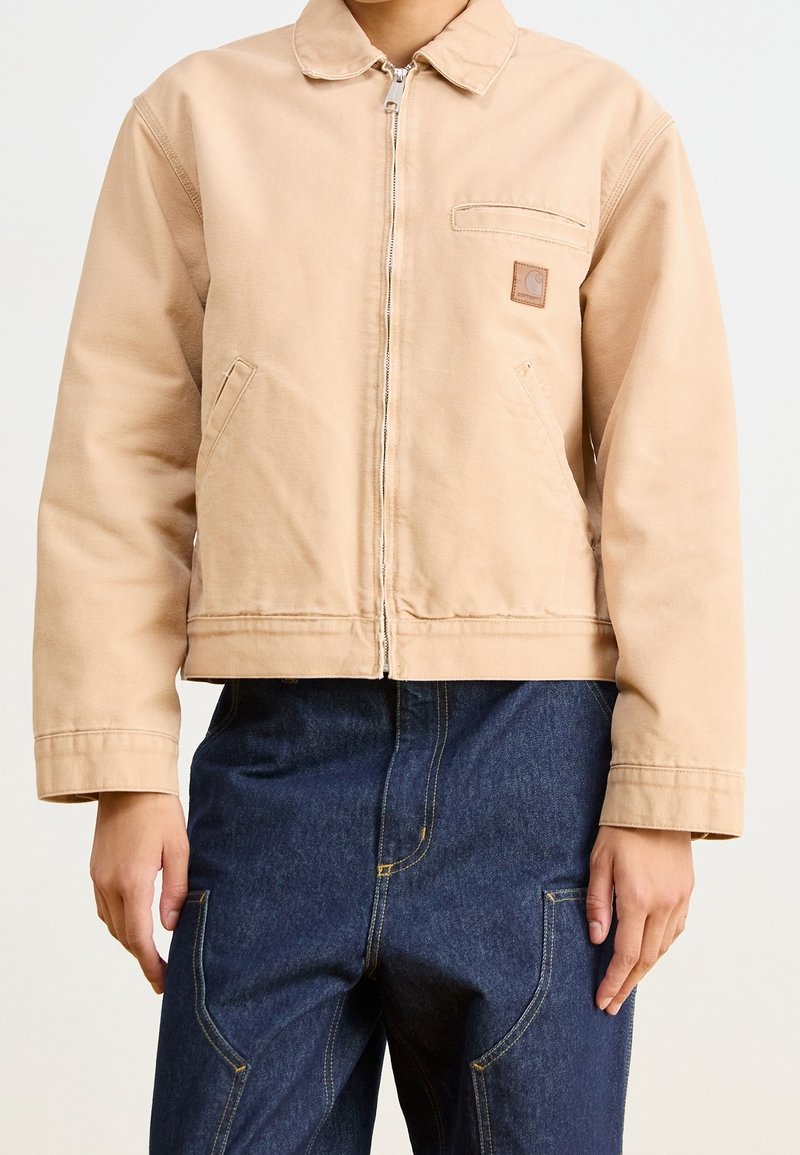 Giacca di cotone beige con zip frontale, colletto piegato e due tasche laterali. Presenta una piccola toppa con il logo sul petto e un design corto.