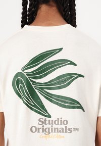Kreminės spalvos marškinėliai su dideliu žaliu lapo piešiniu ir „Studio Originals“ tekstu pilkos ir geltonos spalvos. Trumpomis rankovėmis, laisvas kirpimas.