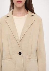 Beige blazer med en strukturerad design, som har en slagkrage, enkel svart knappstängning och två framfickor. Slät tygstruktur.
