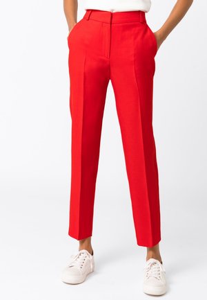 Personne portant un pantalon rouge vif ajusté avec pli marqué à l'avant, poches latérales, et des baskets blanches, debout les mains dans les poches.