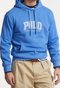 Man med blå Polo Ralph Lauren-hoodie, randig undertröja, beiga byxor och flätad brun bälte, stående med avslappnade händer.