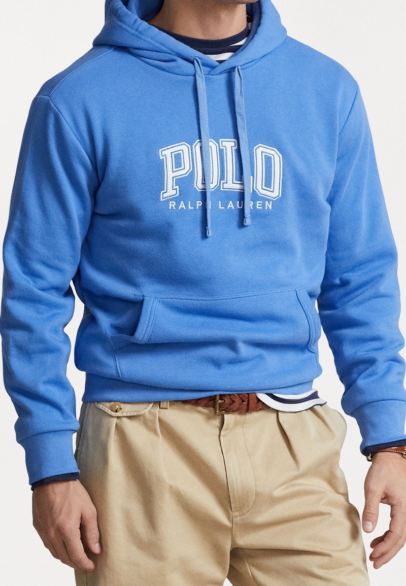 Man med blå Polo Ralph Lauren-hoodie, randig undertröja, beiga byxor och flätad brun bälte, stående med avslappnade händer.