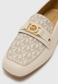 Light beige loafers with a textured fabric upper featuring a subtle "MK" pattern. Gold-tone round logo hardware accents the strap. Rounded toe design.