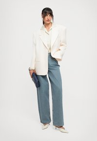 Vrouw in oversized witte blazer, lichte blouse, wijde jeans, witte hakken, met een blauwe clutch tegen een effen achtergrond.
