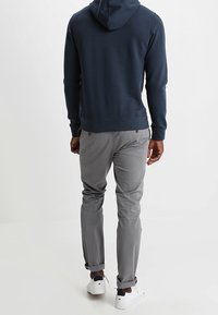 Hombre vestido con sudadera navy, pantalones grises y zapatillas blancas, de pie con la espalda hacia el espectador sobre un fondo claro y liso.