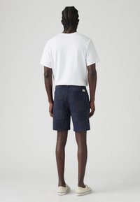 Tricou alb cu mânecă scurtă, pantaloni scurți bleumarin cu motive și puncte deschise la culoare, și încălțăminte de nuanță deschisă. Văzut din spate, arătând detalii ale designului.
