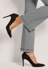 Zwarte suède pumps met hoge hakken, met een slank silhouet en een spitse neus, gecombineerd met wit en zwart gestreepte broek met zigzaglijnen.