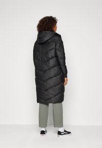 Čierny puffer kabát so vzorom chevron, s kapucňou, dlhými rukávmi a zipsom. V kombinácii so svetlozelenými nohavicami a čiernymi teniskami.
