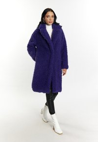 Cămașă din blană artificială violetă, cu guler tăiat și mâneci lungi, asortată cu pantaloni negri mulați și cizme albe cu toc. Textură netedă.