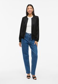 Svart satinbombarjacka med långa ärmar, tillsammans med högmidjade blå denimjeans. Modellen bär svarta klacksandaler.