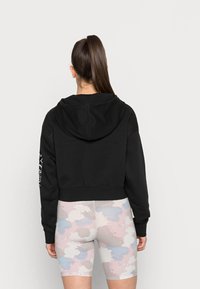Černá zkrácená mikina s kapucí, s texturovaným vzorem a bílou grafikou na levém rukávu, doplněná pastelovými šortkami s maskáčovým vzorem.