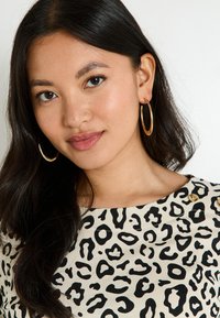 Haut beige avec imprimé léopard noir, doté d'un col rond et de détails en boutons dorés. Le modèle porte des boucles d'oreilles en or et a de longs cheveux foncés.