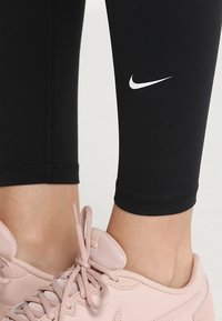 Czarne legginsy wykonane z elastycznego materiału z białym logo Nike na lewej łydce, w połączeniu z jasnoróżowymi butami sportowymi z fakturowanymi detalami.
