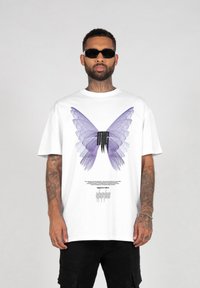 Hvid kortærmet t-shirt med et stort lilla sommerfuglegrafik og sort tekstdesign på forsiden. Modellen bærer sorte solbriller.