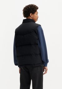 Svart pufferväst med hög krage, prydd med horisontella quiltade sektioner, tillsammans med en blå långärmad skjorta och mörka jeans.