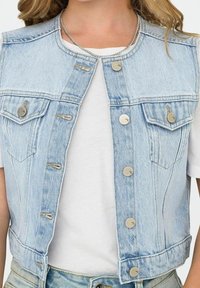 Světle modrá džínová vesta s límcem, pěti kovovými knoflíky a dvěma náprsními kapsami. Nositá přes bílé tričko se surovými okraji.