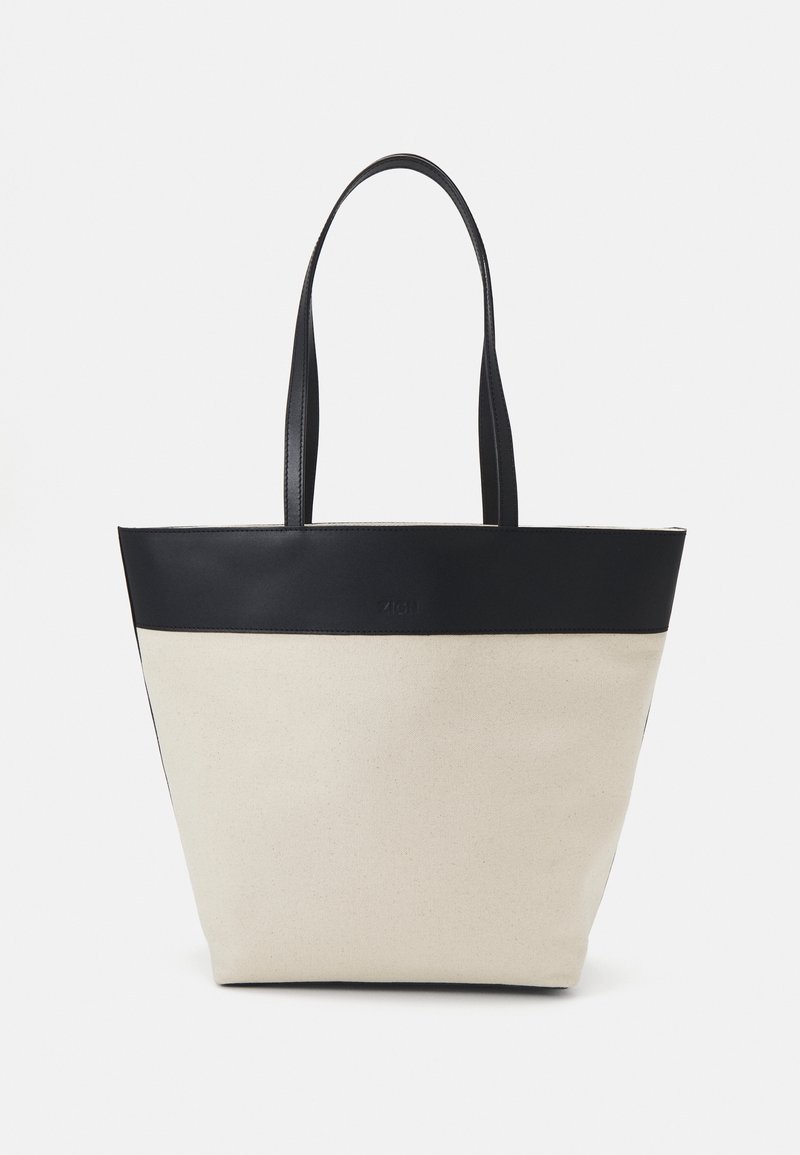 Canvas tote bag with a black leather top panel, two long handles, and a cream-colored body. Features a simple, structured shape.