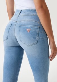Jean taille haute bleu clair avec une coupe slim, présentant un logo de marque rouge triangulaire sur la poche arrière et une texture en denim lisse.