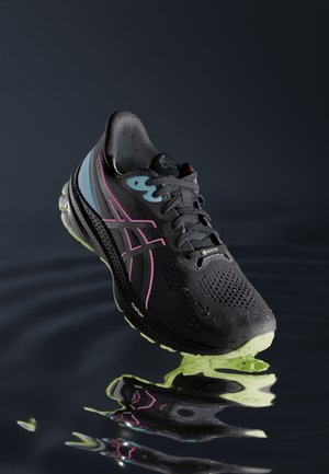 Zapatilla de correr negra y verde neón con acentos rosa y azul, gotas de agua sobre la superficie, reflejada en agua oscura ondulante debajo.