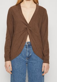 Maglia marrone a maniche lunghe con scollatura a V profonda e dettagli con nodo davanti. Tessuto morbido, leggermente elastico, abbinata a jeans blu a vita alta.