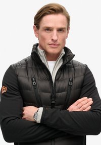 Gilet rembourré noir avec fermeture éclair et doublure en polaire, manches noires, logo orange sur l'épaule gauche, et accents argentés sur une montre.