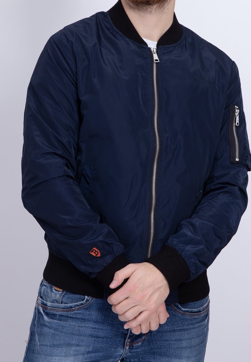 Homme portant une veste bomber bleu marine avec des poignets et un col noirs, fermeture éclair fermée, mains jointes devant, portant un jean bleu.