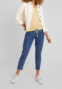 Giacca in velluto a coste bianca con bottoni frontali, blusa gialla sotto e jeans blu a sigaretta. Abbinata a sneakers bianche, con maniche arrotolate.