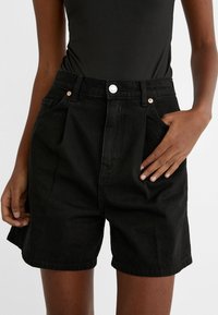 Shorts de mezclilla negra con talle alto, con pliegues, bolsillos frontales y herrajes plateados. Textura suave y diseño a la altura de la rodilla.