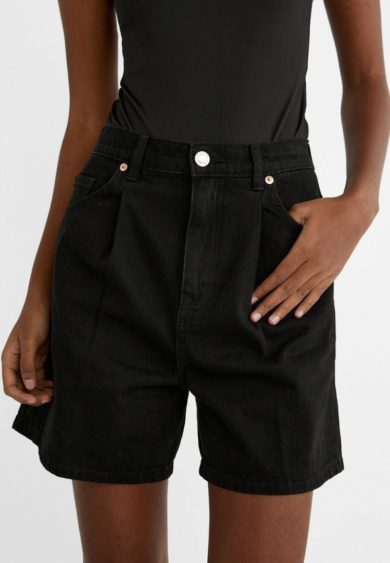 Shorts de mezclilla negra con talle alto, con pliegues, bolsillos frontales y herrajes plateados. Textura suave y diseño a la altura de la rodilla.