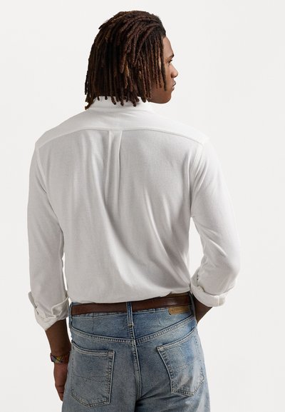White long-sleeve shirt with a relaxed fit, featuring a back yoke seam and rolled-up cuffs, paired with light blue denim jeans.
