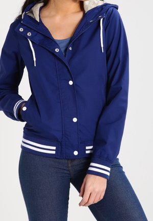 Donna che indossa una giacca con cappuccio blu con laccetti bianchi, bottoni a pressione, polsini e orlo a righe, abbinata a jeans blu scuro.