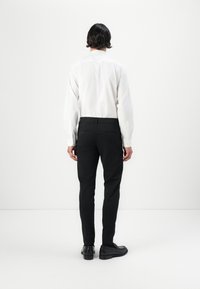 Weißes Langarmhemd kombiniert mit schwarzen maßgeschneiderten Hosen und schwarzen Schuhen, von hinten vor einem schlichten weißen Hintergrund betrachtet.