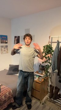 Noor mees halli särke ja teksadega tõstab mõlemaid käsi magamistoas, kus on voodi, taim, riidepuu ja öökapp raamatute ning laualambiga.