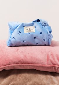Manta de forro polar azul con pequeños patrones de lazo azul y una etiqueta que dice "THE FLAT LAY CO." sobre un cojín texturizado rosa.