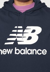 Henkilö pukeutuneena tummansiniseen huppariin, jossa valkoinen New Balancen logo ja teksti, päällä violetti pyöreäkauluksinen paita.