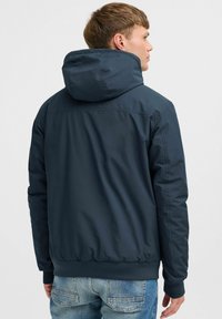 Chaqueta con capucha de color azul marino con un acabado suave, puños elásticos y dobladillo. La parte trasera presenta un diseño simple y aerodinámico sin hardware visible.