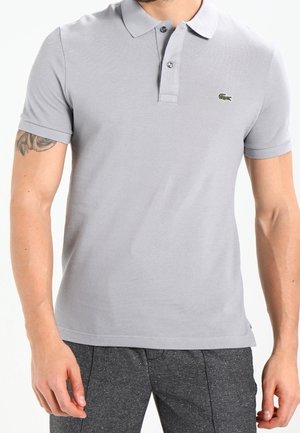 Mann trägt ein hellgraues Polohemd mit kurzen Ärmeln, Kragen und drei Knöpfen, kombiniert mit dunkelgrauen Hosen.