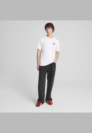 T-shirt bianco in cotone con una piccola grafica blu sul petto, abbinato a pantaloni in denim neri larghi e scarpe rosse.