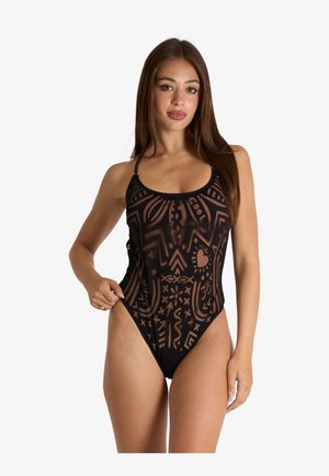 Donna con lunghi capelli castani che indossa un costume intero nero trasparente con motivi astratti, in piedi davanti a uno sfondo chiaro uniforme.