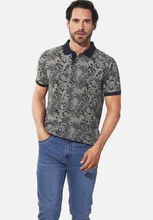 Poloshirt mit Blumenmuster in Olivgrün, ausgestattet mit einem marineblauen Kragen und Ärmelsäunen, kombiniert mit blauen Jeans und in einem lässigen Schnitt.