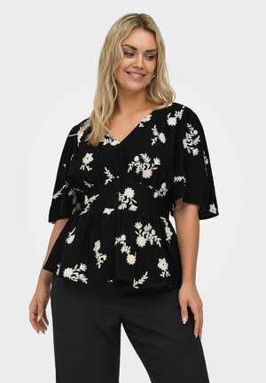 Kvinde iført sort bluse med hvid blomsterbroderi og korte ærmer, kombineret med sorte bukser, smilende og kiggende en smule mod venstre.