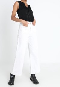 Mustaton hihaton toppi V-kauluksella, yhdistettynä korkeavyötäröisiin valkoisiin leveälahkeisiin farkkuihin ja mustiin nilkkuriin. Yksinkertainen, siisti muotoilu.