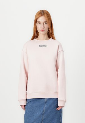 Light pink sweatshirt with a round neckline and dropped shoulders, featuring a stitched "LACOSTE" logo on the chest. Worn over a denim skirt.