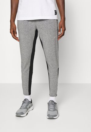 Man wearing grey athletic pants with black side panels, white socks, grey sneakers, and a white T-shirt standing against a plain background.