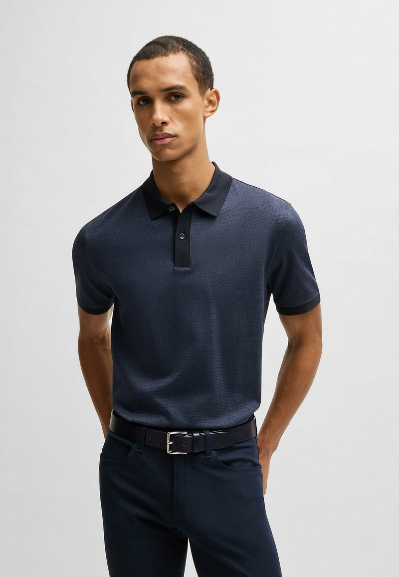 Polo shirt blu navy con motivo testurizzato, colletto nero e patta con due bottoni. Abbinato a pantaloni scuri e una cintura nera.